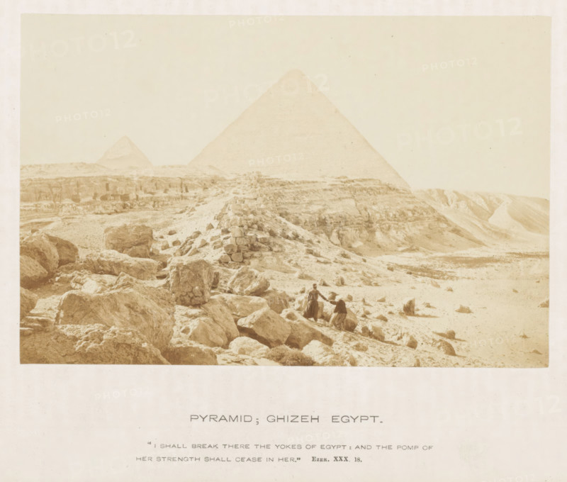 Pyramid, Giza Egypt, View of the Pyramid of Cheops, photograph, page, inscription: ''I shall break there the yokes of Egypt: and the pump of her strength shall cease in here.' Ezek. XXX. 18.', photographer: Francis Frith (possibly), Giza, c. 1850 - c. 1865, North Africa, paper, albumen print, height 104 mm x width 157 mm, Egyptian pyramids (Wonder of the World), Pyramids of Giza, A sepia-toned photograph shows a desert landscape with large stone blocks and rocky terrain in the foreground leading toward two pyramids in the background, one pyramid larger and closer and the other smaller and more distant, with scattered rubble and a low stone cairn or mound on the slope; two figures in traditional clothing stand among the rocks near the center, one standing and one seated, with undulating sandy slopes and eroded cliffs visible to the right and a pale sky filling the upper portion of the image.