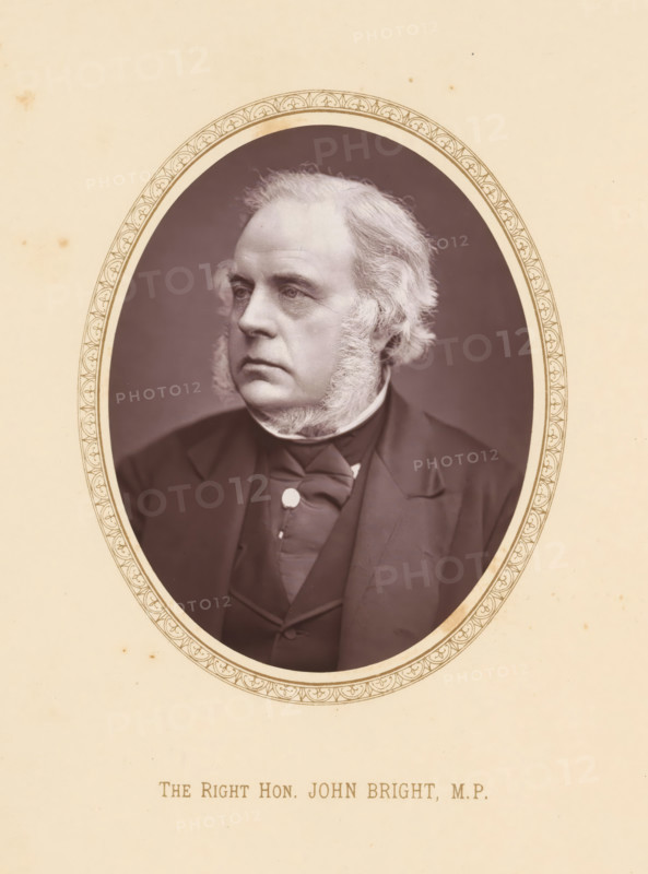William Ewart Gladstone, page, photomechanical print, photographer: John Moffat, Edinburgh, c. 1875 - c. 1880, printmaker, paper, Woodburytype, height 109 mm x width 81 mm, Person, politician, eg party leader, An oval-framed sepia portrait of an older man shown from the chest up, wearing a dark suit jacket, waistcoat, and a bow or cravat at the neck, with prominent sideburns and light-colored hair swept back, his face turned slightly to one side and set against a dark oval background surrounded by a decorative border on a pale page.