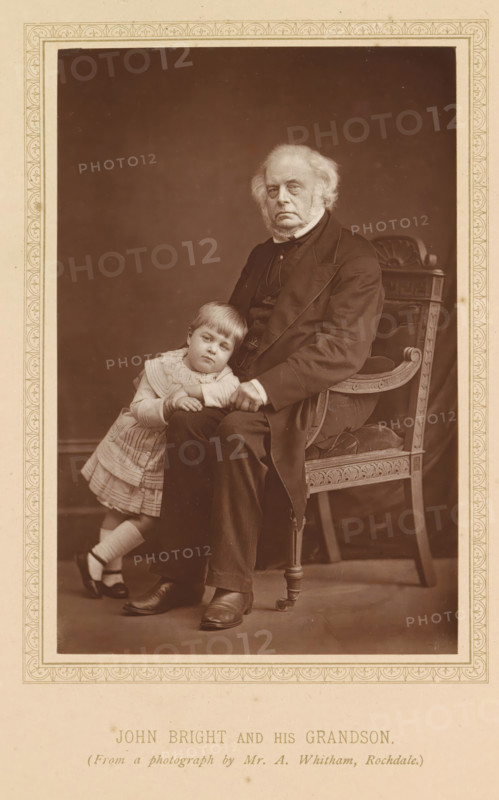 Portrait of John Bright and his grandson, John Bright and his grandson, page, photomechanical print, photographer: A. Whitham, Rochdale, c. 1874 - in or before 1884, printmaker, Manchester, paper, Woodburytype, Woodburytypepie, height 144 mm x width 95 mm, Print after title page., Person, John Bright, grandparent(s) with grandchild(ren) (family group), An older man with white hair and sideburns sits on an ornate wooden chair with carved details and upholstered seat, wearing a dark suit and shoes, while a young child in a light dress, socks, and dark shoes leans against his knee and rests their hands on his leg, both posed against a plain dark backdrop within a decorative rectangular border.