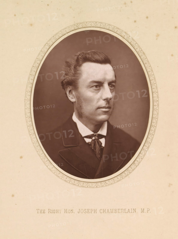 Portrait of Joseph Chamberlain, The right hon. Joseph Chamberlain, MP, page, photomechanical print, c. 1874 - in or before 1884, photographer, printmaker, paper, Woodburytype, height 111 mm x width 87 mm, Person, Joseph Chamberlain, politician, eg party leader, A sepia-toned oval portrait of a clean-shaven man with sideburns and neatly combed hair, shown from the shoulders up and turned slightly to his left, wearing a dark suit jacket, a white shirt with a high collar, and a patterned cravat, all set against a plain dark background and mounted on a textured light-colored mat with a decorative oval border.