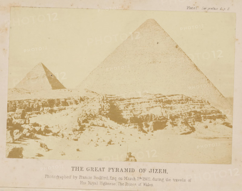 The great pyramid of Jiza, photograph, page, inscription: 'Photographed by, Prince of Wales', number: 'Plate 1.', photographer: Francis Bedford, Giza, 1862-03-05, North Africa, paper, albumen print, albumendruk, height 137 mm x width 92 mm, Photo on frontispiece., Egyptian pyramids (Wonder of the World), Pyramid of Cheops, A faded sepia photograph showing a desert scene with two large pyramidal stone structures rising from a rocky, uneven foreground; the larger pyramid is on the right and the smaller pyramid is to the left with a triangular darker face visible, while the foreground displays scattered stones, ledges, and eroded rock formations under a pale sky.