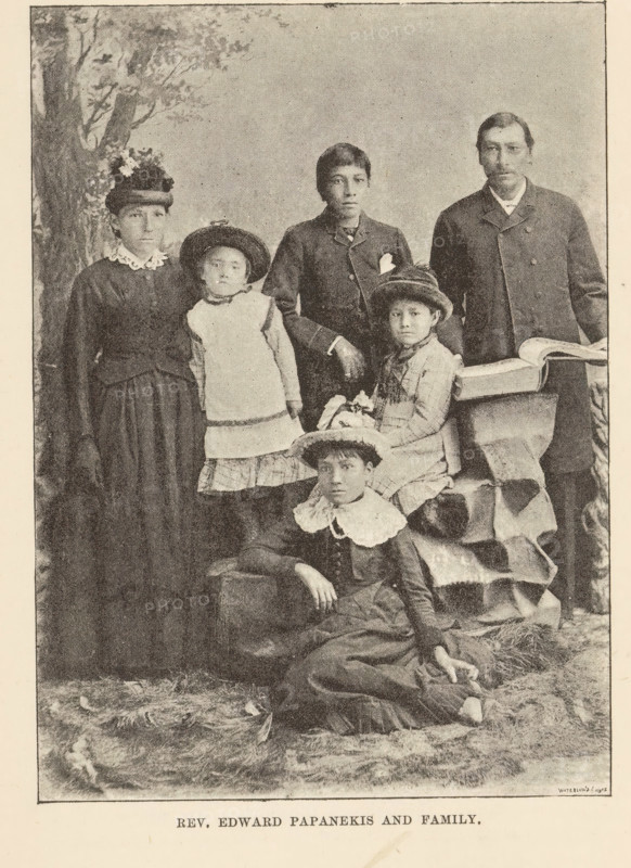 Portrait of Edward Paupanekis and his family, Rev. Edward Papanekis and family, page, photomechanical print, Canada, maker: Waterlow & Sons Limited, c. 1882 - in or before 1892, photographer, North America, paper, autotypie, height 135 mm x width 97 mm, Person, Edward Paupanekis, human races, peoples, nationalities, family, descent, A sepia-toned vintage studio photograph showing a group of people arranged in formal poses; a standing woman on the left wears a dark dress with a high lace collar and a hat decorated with flowers, next to a young girl in a light pinafore and a round hat leaning slightly; a standing boy in a dark jacket with a hand resting on a seated girl's shoulder stands center, the seated girl wearing a dress with a small hat and holding a small object in her lap; a man in a dark coat stands on the right beside a draped table with an open book on it; in front a young woman reclines on the floor wearing a dress with a large white collar and a hat, one arm resting on a ra...