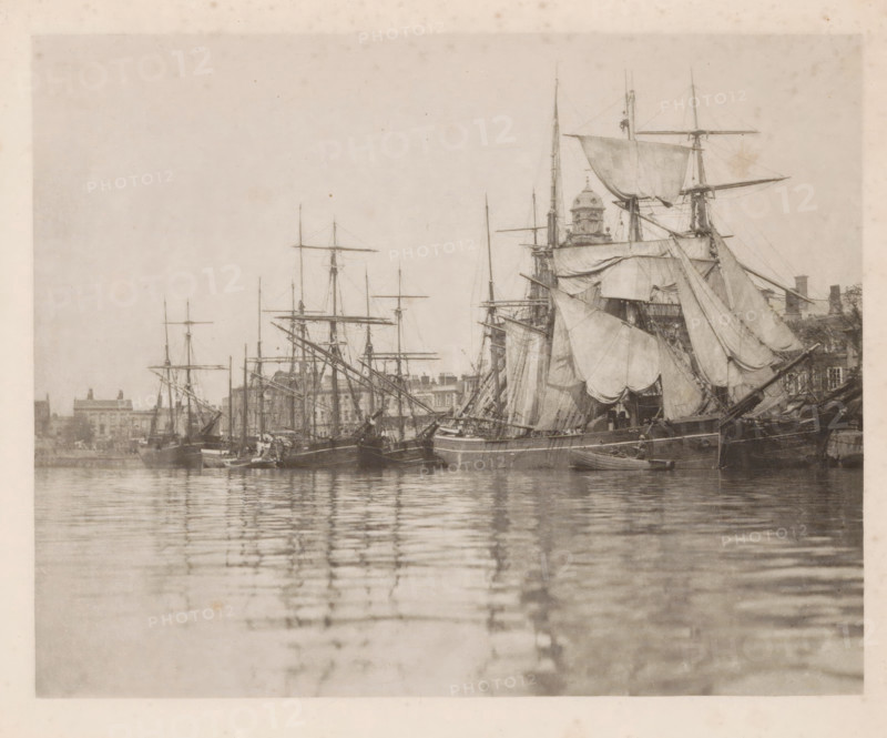 Sailing ships in the harbor of Great Yarmouth, page, photomechanical print, title: 'Great Yarmouth harbour', number: 'Plate I', photographer: Peter Henry Emerson, Great Yarmouth, cliché maker: Walter L. Colls, c. 1880 - in or before 1890, Norfolk (county), paper, photoaquatint, height 177 mm x width 221 mm, Print on frontispiece., harbour, ships (in general), A sepia-toned photograph showing a harbor scene with multiple sailing ships moored closely together, tall masts and rigging rising against a pale sky, large furled and partially unfurled sails draped across the vessels, a prominent ship in the foreground with heavy canvas and wooden hull details, smaller boats alongside, calm water in the foreground reflecting the masts and hulls, and a low line of buildings and architectural elements visible along the distant shoreline behind the ships.