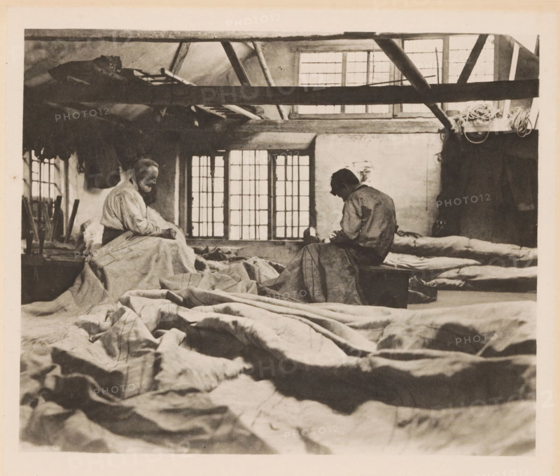 Interior of a sail-making workshop, page, photomechanical print, number: 'Plate XVIII', inscription: 'In a sail-loft', photographer: Peter Henry Emerson, cliché maker: Walter L. Colls, c. 1880 - in or before 1890, paper, photoaquatint, height 164 mm x width 199 mm, interior, representation of a building, sail, A sepia-toned interior scene showing two seated figures facing each other on low surfaces with large rumpled sheets and fabric spread across the foreground and floor, exposed ceiling beams and rafters crossing the top of the image, a row of mullioned windows behind the figures letting diffuse light into the room, coiled ropes hanging near the upper right, a variety of folded and stacked bedding along the right wall, and a cluttered work area with tools and garments visible at the left.