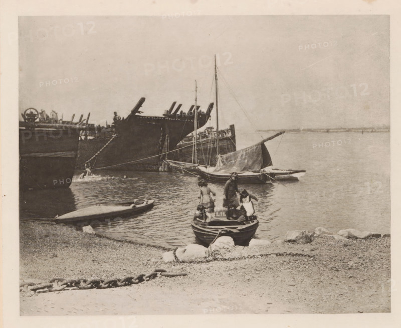 Children in a rowboat on the Breydon, page, photomechanical print, number: 'Plate IV', title: 'Old hulks', photographer: Peter Henry Emerson, Breydon, cliché maker: Walter L. Colls, c. 1880 - in or before 1890, Norfolk (county), paper, photoaquatint, height 161 mm x width 201 mm, rowing-boat, canoe, child, ships (in general), A shoreline scene with small rowboats and larger sailing vessels moored near the water, a small boat pulled up on the pebbled beach, a larger boat in the foreground with people standing and sitting inside it, a sailboat with its sail furled floating nearby, taller ships with masts and rigging visible further out, calm water extending toward the horizon, a chain lying on the beach in the foreground, scattered rocks along the water's edge, and an overcast sky above.