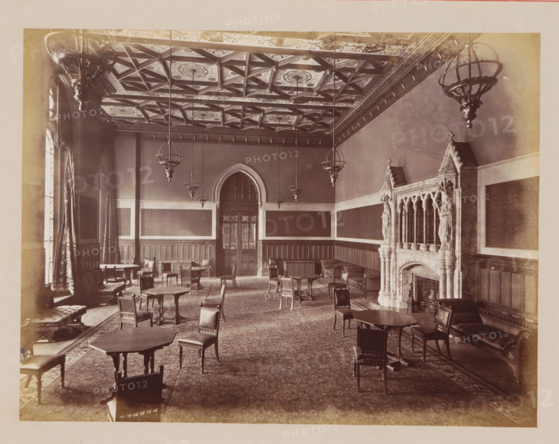 Reception Room in Manchester City Hall, photograph, page, title: 'The Reception-Room', photographer: J. McLeod, Manchester City Hall, 1877, Manchester, paper, albumen print, height 162 mm x width 214 mm, town hall, interior, representation of a building, A large ornate room with an intricately patterned coffered ceiling and multiple hanging spherical chandeliers, wood-paneled walls with tall rectangular panels, a central arched doorway with glass panes, numerous wooden tables and chairs scattered across a patterned carpet, tall draped windows along the left wall, cushioned benches and armchairs along the sides, and an ornate carved stone fireplace with decorative columns and statues on the right wall.