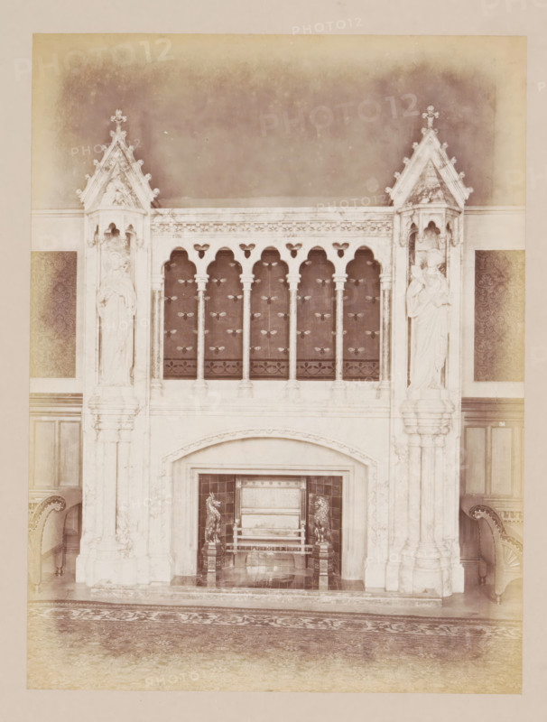 Fireplace in the reception room in Manchester Town Hall, photograph, page, title: 'The Reception-Room Chimney-Piece', photographer: J. McLeod, Manchester Town Hall, 1877, Manchester, paper, albumen print, height 200 mm x width 151 mm, town hall, interior, representation of a building, chimney-piece, mantelpiece, A tall ornate stone fireplace with carved pillars and gothic arches occupies the center of the image, flanked on each side by sculpted figures set into pointed canopied niches; above the fireplace is a decorative balustrade with slender columns and a patterned dark panel behind them, and below the arch of the fireplace opening is an ornate hearth area containing a bench or seat with sculpted animal figures at its sides; portions of upholstered chairs are visible at the left and right edges, and a patterned rug covers the floor in front of the fireplace.