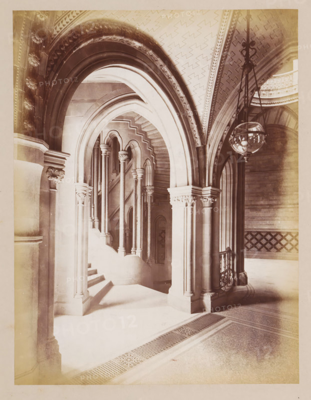 Staircase in Manchester Town Hall, photograph, page, title: 'The Circular Staircases', photographer: J. McLeod, Manchester Town Hall, 1877, Manchester, paper, albumen print, height 213 mm x width 162 mm, townhall, staircase, interior, representation of a building, A sepia-toned interior scene showing a series of ornate stone arches and columns leading to a curved staircase, with multiple slender columns topped by decorative capitals and pointed archways visible through successive openings, a patterned vaulted ceiling with geometric motifs above, a suspended glass lantern hanging from ornate metal chains on the right, carved floral and foliate details on column capitals and arch mouldings, a decorative railing beside the staircase, and a tiled floor with a perforated grate and patterned border in the foreground.