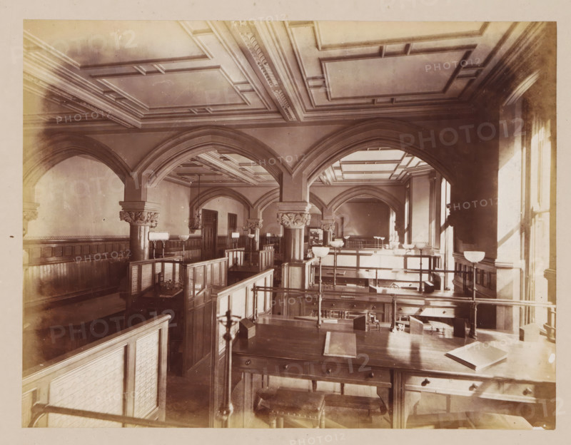 Office in Manchester Town Hall, photograph, page, title: 'The Gas Office', photographer: J. McLeod, Manchester Town Hall, 1877, Manchester, paper, albumen print, height 162 mm x width 213 mm, office (, administration), town hall, interior, representation of a building, A large interior space with high decorative ceilings and multiple arched openings supported by columns with carved capitals, wooden paneling along the walls, rows of wooden desks and partitions forming cubicles, several tall lamps with bowl-shaped shades mounted on stands, a broad wooden table in the foreground with drawers and a closed book resting on it, railings and stair-like platforms dividing levels, tall windows on the right casting light and shadows across the room, and ornamental moldings and recessed panels on the ceiling.