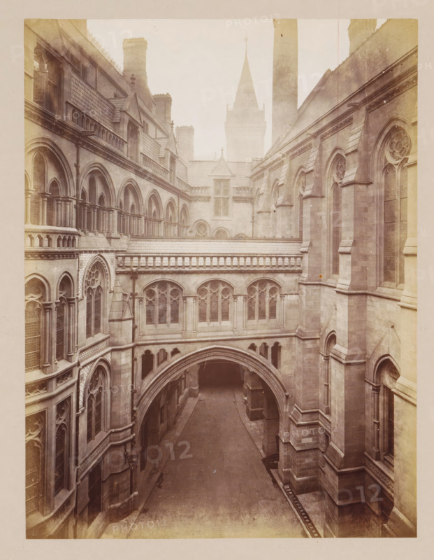 Courtyard of Manchester City Hall, photograph, page, title: 'Princess Street Courtyard and Bridge', photographer: J. McLeod, Manchester City Hall, 1877, Manchester, paper, albumen print, height 213 mm x width 160 mm, courtyard, townhall, A narrow stone courtyard flanked by tall gothic buildings with arched windows and decorative tracery, featuring a covered walkway with a series of small columns and balustrades above an arched passageway, steep slate roofs with dormer windows and chimneys, a pointed tower rising in the background, paved ground below the arch, and weathered stone facades with carved moldings and buttresses.
