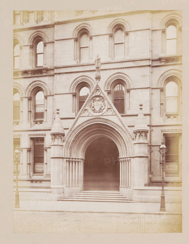 Entrance portal of Manchester City Hall, photograph, page, title: 'Cooper Street Entrance', photographer: J. McLeod, Manchester, 1877, paper, albumen print, height 213 mm x width 162 mm, town hall, façade (of house or building), gate, entrance, Manchester City Hall, A stone building facade with a large arched entrance framed by clustered columns and a triangular pediment topped by a standing statue, flanked by ornate pinnacles and carved decorative elements, multiple arched windows with recessed frames across two visible stories, stone steps leading up into the dark archway, patterned cobblestone street in front, and two tall street lamps positioned near the entrance.