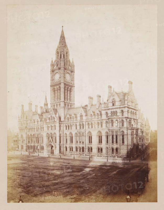 Exterior of Manchester Town Hall, photograph, page, photographer: J. McLeod, Manchester, 1877, paper, albumen print, height 212 mm x width 162 mm, town hall, façade (of house or building), Manchester Town Hall, A large stone building with ornate Gothic architecture fills the frame, featuring a tall central tower with a spire, clock faces, arched windows, decorative turrets, and multiple chimneys; the façade shows rows of arched windows, carved stone details, and an arched main entrance; a wide paved street in front of the building appears worn and wet, bordered by sidewalks and lampposts; the overall image has a faded, sepia-toned appearance.