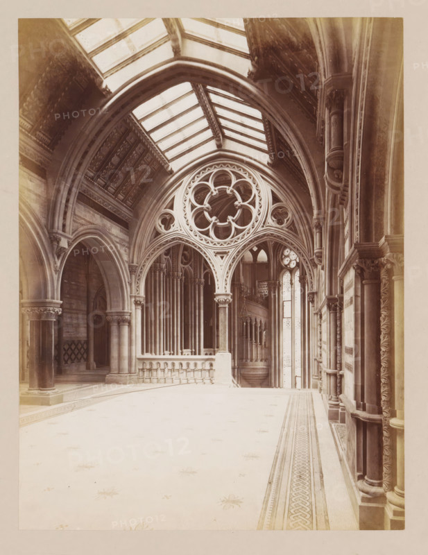 Vestibule in Manchester City Hall, photograph, page, title: 'The Ante-Hall', photographer: J. McLeod, Manchester City Hall, 1877, Manchester, paper, albumen print, height 202 mm x width 152 mm, vestibule, hall, townhall, A vaulted interior space with tall arches and columns, featuring an ornate circular tracery window set above a carved archway, a row of slender clustered columns behind a low balustrade, patterned floor with a decorative border, a high glass-panelled ceiling, carved capitals and mouldings along the walls, and tall narrow windows with decorative glazing in the background.