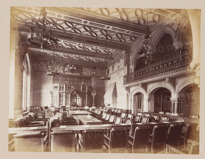 Meeting room in Manchester City Hall, photograph, page, title: 'The Council Chamber', photographer: J. McLeod, Manchester City Hall, 1877, Manchester, paper, albumen print, height 162 mm x width 204 mm, townhall, interior, representation of a building, provincial government, A large ornate hall with a patterned coffered ceiling and multiple hanging chandeliers, rows of leather-upholstered chairs arranged around long wooden tables, tall arched windows along one wall, a carved stone fireplace with a stepped hood, a wooden carved screen or gallery at the far end, decorative wall panels and friezes above the paneling, an elevated balcony with an intricate metal railing supported by stone arches and columns, and various lamps and candlesticks placed throughout the room.