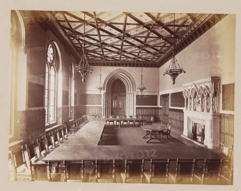 Mayor's Parlour in Manchester City Hall, photograph, page, title: 'The Mayor's Parlour', photographer: J. McLeod, Manchester City Hall, 1877, Manchester, paper, albumen print, height 161 mm x width 212 mm, town hall, interior, representation of a building, A large rectangular room with a patterned coffered ceiling featuring diagonal beams and decorative panels, three ornate hanging chandeliers suspended from the ceiling, tall arched windows along one long wall with framed panels beneath them, long wooden tables arranged in a U shape surrounded by high-backed wooden chairs, an arched doorway with layered stone molding centered on the far wall, dark wooden wainscoting and wall panels lining the room, an elaborately carved stone fireplace with clustered columns and decorative arches on the right wall, a smaller standalone table and chairs near the fireplace, and a patterned floor covering the central area.