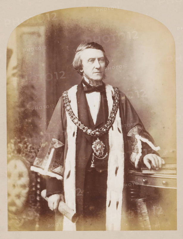 Manchester town hall, book, inscription: 'In commemoration, Hall Sub-Committee.', inscription: 'ex libris SFJ', photographer: J. McLeod, 1877, plant material, biological material, paper, wood (plant material), leather, printing, albumen print, color on cut, height 330 mm x width 275 mm x thickness 42 mm, A sepia-toned portrait of a middle-aged man standing and facing forward, wearing a dark formal coat with a high collar and a bow tie, layered with a fur-trimmed ceremonial robe and an ornate chain of office draped around his shoulders; his hair is parted to one side and he has distinctive sideburns and a short beard on his chin, his left hand rests on a wooden table with carved details while his right hand holds a rolled document, and the background shows a softly blurred interior with mottled tonal variations.