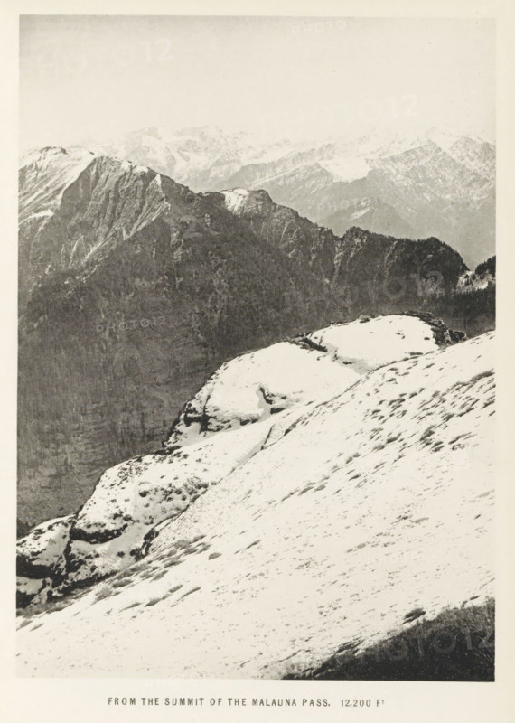 View from the summit of the Malana pass, Himachal Pradesh, India, page, photomechanical print, caption: 'FROM THE SUMMIT OF THE MALAUNA PASS. 12, 200 Ft', photographer: Frederick Saint John Gore, Malana, c. 1890 - in or before 1895, printmaker, Parvati valley, paper, collotype, photoprint, height 179 mm x width 129 mm, mountain-range, snow peaks, A snowy mountain slope fills the foreground with patches of exposed rock and sparse vegetation, leading to rounded snow-covered ledges and rocky outcrops; behind them a darker, steep ridgeline rises with craggy cliffs and forested sections, and in the distant background a range of rugged, snow-dusted peaks recedes under a pale sky.