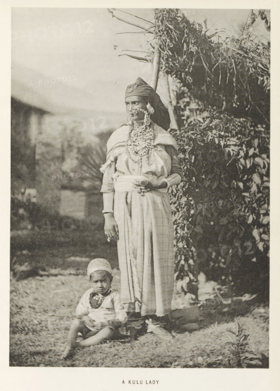 Woman with a child at Kullu, Himachal Pradesh, India, page, photomechanical print, caption: 'A KULU LADY', photographer: Frederick Saint John Gore, Kullu, c. 1890 - in or before 1895, printmaker, Kullu Valley, paper, collotype, photoprint, height 179 mm x width 129 mm, adult woman, child, ornaments, jewels, A sepia-toned photograph of a standing woman and a seated child outdoors, the woman wearing a headwrap, layered necklaces and facial jewelry, a shawl draped over her shoulders and a long patterned skirt, her hands resting at her waist with one holding an object, and the child seated on the ground in front of her wearing a cap and simple clothing; foliage and a thatched structure appear in the background.