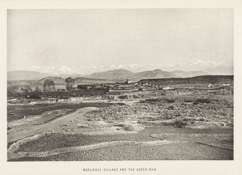View of Maru Khel, Khyber Pakhtunkhwa, Pakistan, page, photomechanical print, caption: 'MARUKHEL VILLAGE AND THE SAFED KÓH', photographer: Frederick Saint John Gore, Maru Khel, c. 1890 - in or before 1895, printmaker, paper, collotype, photoprint, height 120 mm x width 178 mm, city-view in general, 'veduta', mountain-range, snow peaks, A wide landscape photograph showing a foreground of rough, rocky ground with visible furrows and a winding dirt path, leading back to a middle ground of low, flat agricultural terraces and scattered trees, a small cluster of low buildings and structures set against gently rolling hills, and a distant range of higher mountains with snow-capped peaks under a broad, pale sky.