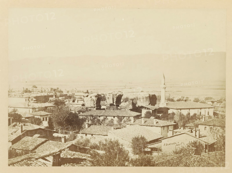 View of Alasehir, photograph, page, photographer: Alexander Svoboda, Alasehir, c. 1859 - in or before 1869, Ottoman Empire, paper, albumen print, height 128 mm x width 177 mm, Photo for page 35., ruin of a building, architecture, A sepia-toned panoramic view of a town with clustered tiled rooftops in the foreground, scattered trees and vegetation among the buildings, a mosque with a slender minaret rising above the structures toward the right, several ruined stone arches or large rock formations near the center, low-lying fields stretching into the distance, and a faint silhouette of a mountain ridge along the horizon under a pale sky.