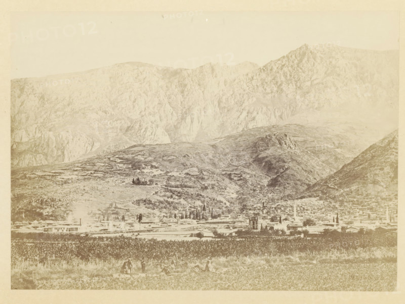 View of the ruins of Magnesia on the Sipylos, photograph, page, photographer: Alexander Svoboda, Asia Minor, c. 1859 - in or before 1869, paper, albumen print, height 129 mm x width 179 mm, ruin of a building, architecture, (high) hill, Magnesia on the Sipylos, A wide landscape photograph showing a mountainous backdrop with steep rocky slopes and ridges, a midground of rolling hills with scattered trees and terraced areas, and a foreground containing a flat field with rows of vegetation and a few small groups of people standing or sitting; along the base of the hills there is a spread of low buildings and a taller tower-like structure among them, and the overall image has a faded, sepia-toned appearance.