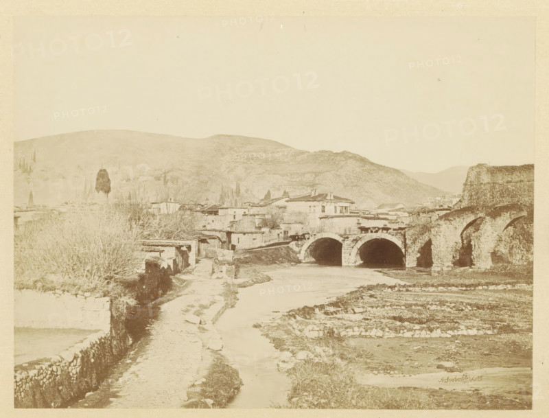 View of Pergamon, photograph, page, photographer: Alexander Svoboda, Pergamon, c. 1859 - in or before 1869, Asia Minor, paper, albumen print, height 124 mm x width 176 mm, city-view in general, 'veduta', bridge in city across river, canal, A sepia-toned landscape showing a river running toward the foreground with a dirt path alongside stone walls, a stone arched bridge crossing the river with multiple arches, adjoining stone structures and low buildings clustered near the bridge, a taller building with a tiled roof near the center, leafless shrubs and trees along the banks, a large rounded hill rising in the background, and expanses of sky above.