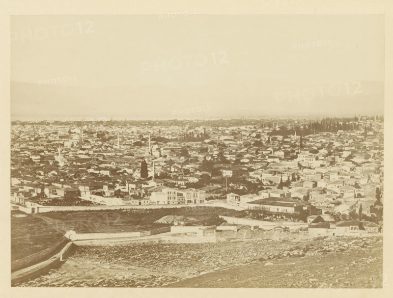 View of Smyrna, photograph, page, photographer: Alexander Svoboda, Smyrna, c. 1859 - in or before 1869, Ottoman Empire, paper, albumen print, height 130 mm x width 178 mm, city-view in general, 'veduta', A wide sepia-toned panoramic view showing an expansive cityscape with densely packed low-rise buildings and varied rooflines, several slender vertical towers rising among the structures, clusters of tall trees interspersed through the urban area, a low stone wall running across the foreground above a textured, rocky slope, and a pale sky occupying the upper portion of the image.
