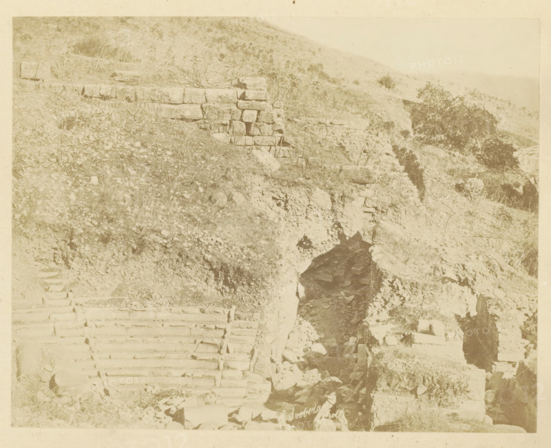 The Odeon of Ephesus, photograph, page, photographer: Alexander Svoboda, Ephesus, c. 1859 - in or before 1869, Efeze, paper, albumen print, height 139 mm x width 177 mm, ruin of a building, architecture, A sepia-toned scene showing a rocky hillside with irregular stone steps carved into the slope and a rough stone retaining wall partially visible, scattered rocks and sparse grasses covering much of the terrain, a narrow dark crevice or opening in the rock face, and low vegetation and small shrubs dotting the hillside.
