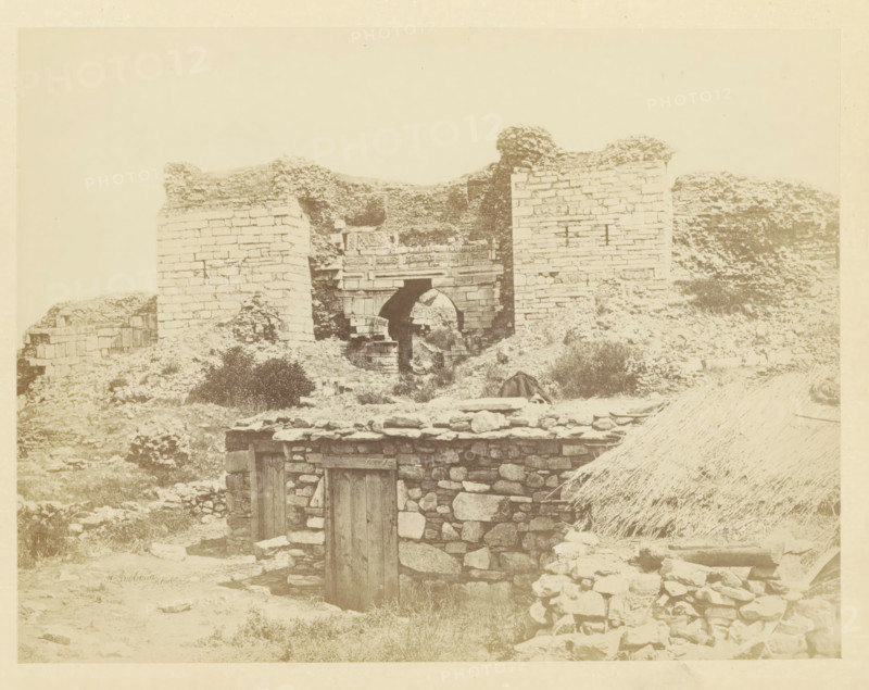 View of the Gate of Persecution on the hill of Ayasoluk, photograph, page, photographer: Alexander Svoboda, Asia Minor, c. 1859 - in or before 1869, paper, albumen print, height 134 mm x width 174 mm, gate, entrance, A sepia-toned photograph showing stone ruins on uneven ground with a partially collapsed arched gateway set between two tall masonry walls, rubble and low stone structures in front of the gateway, a small stone building with a wooden door in the foreground, scattered rocks and vegetation around the site, and a thatched roof visible at the lower right edge.