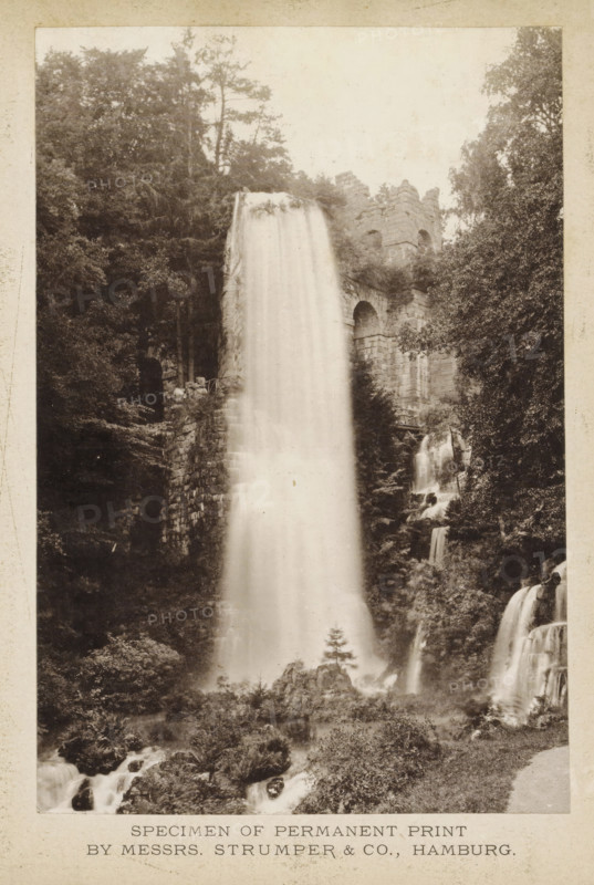 Waterfall, page, photomechanical print, inscription: 'Specimen of permanent print by Messrs. Strumper & Co., Hamburg', photographer: Strumper & Co (possibly), Hamburg (city), c. 1868 - in or before 1878, paper, collotype, photoprint, height 158 mm x width 107 mm, A tall, cascading waterfall flows down a rocky cliff surrounded by dense trees and foliage, with water streaming smoothly from the top and breaking into smaller falls along the right side; stone ruins with arched openings and blockwork sit near the upper falls, partially covered by vegetation, and rocky outcrops and plants appear at the base where mist rises from the falling water.