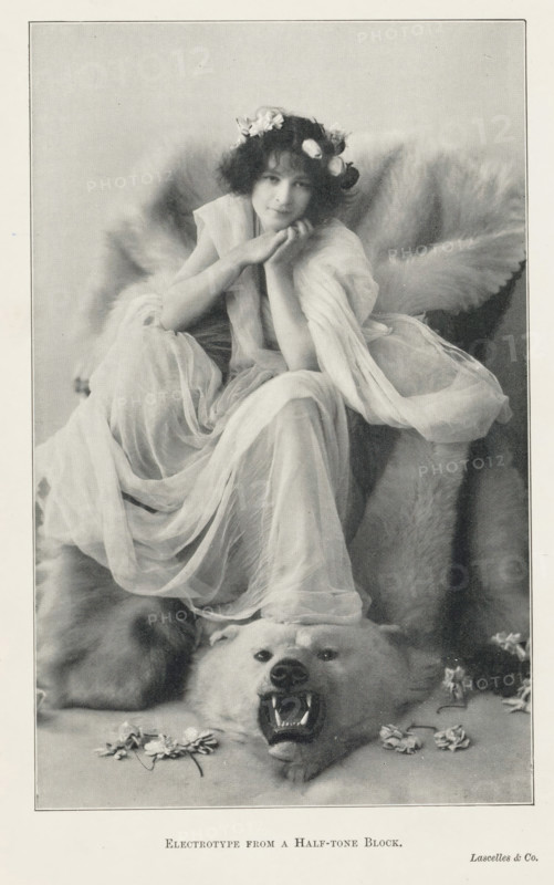 Portrait of an unknown woman on a polar bear skin, page, photomechanical print, printmaker: Lascelles & Co., c. 1889 - in or before 1899, photographer, paper, autotype, electrotype from a half-tone block, height 176 mm x width 111 mm, adult woman, beasts of prey, predatory animals: polar bear, A seated young woman with a wreath of flowers in her hair rests her chin on her hands, wearing a flowing translucent gown that drapes around her and cascades over a large fur beneath her; behind her the fur forms a voluminous backdrop, and at her feet a bear skin with an open mouth and visible teeth lies flat on the ground, with scattered petals and small flowers near the bear’s head.