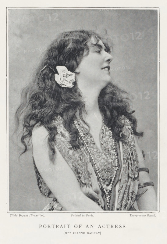 Portrait of an actress, page, photomechanical print, photographer: Louis Désiré Dupont, Brussels, printmaker: Goupil & Cie., c. 1889 - in or before 1899, paper, autotypie, typogravure, height 133 mm x width 98 mm, Person, Jeanne Raunay, portrait of actor, actress, A black and white portrait of a woman shown from the waist up in profile facing right, with long wavy hair cascading over her shoulders and a large flower tucked behind one ear, wearing a patterned sleeveless garment and multiple layered necklaces, her head tilted slightly back and her lips parted in a slight smile, framed by a rectangular border and a light textured backdrop.