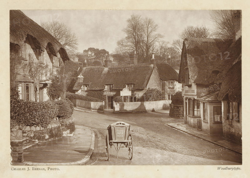 Crab Inn, Shanklin, Isle of Wight, page, photomechanical print, photographer: Charles J. Brenan, c. 1889 - in or before 1899, printmaker, paper, Woodburytype, Woodburytypepie, height 98 mm x width 145 mm, inn, coffee-house, public house, street, A sepia-toned street scene shows a narrow road curving past thatched cottages with low, textured roofs and small multi-paned windows, flanked by hedges and stone walls. A simple two-wheeled cart is positioned on the road in the foreground, viewed from behind. On the left, a paved footpath runs beside a stone wall topped by trimmed bushes and leads toward a row of cottages partly covered in climbing plants. In the center, a fenced entrance opens to a cluster of cottages with a lamp post nearby and chimney stacks rising above the thatch. On the right, another thatched building presents a projecting bay window and timbered details, with bare trees visible behind the rooftops and additional houses set further back on a gentle rise. The sky appears ...