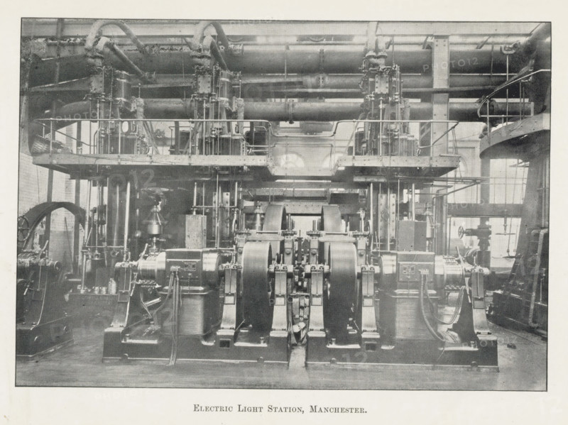 Electric light stations, Manchester, page, photomechanical print, photographer: Brothers, WJ, c. 1889 - in or before 1899, printmaker, paper, autotypie, height 109 mm x width 157 mm, power station, exterior, representation of a building, A wide interior view of an industrial room filled with large metal machinery and piping, showing multiple heavy cylindrical flywheels and curved belts in the foreground mounted on sturdy bases, a raised platform with railings and vertical supports running across the midsection, complex networks of pipes and fittings overhead and behind the platform, structural columns and beams framing the space, various valves, gears, and smaller mechanical components scattered around the floor level, and tall windows at the rear letting in light.