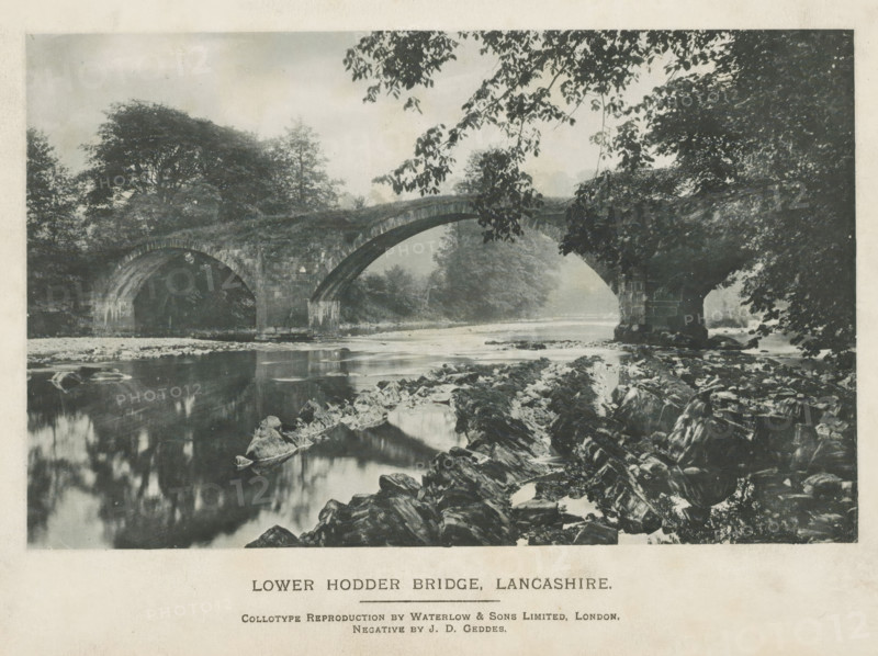 Lower Hodder Bridge, Lancashire, page, photomechanical print, photographer: JD Geddes, printmaker: Waterlow & Sons Limited, c. 1889 - in or before 1899, paper, collotype, photoprint, height 101 mm x width 161 mm, landscape with bridge, viaduct or aqueduct, A monochrome scene showing a stone arched bridge spanning a calm river, with two large arches visible and a rocky riverbank in the foreground; overhanging tree branches frame the top and right edges, while dense trees and foliage fill the background on both sides of the bridge and reflect faintly in the water below.