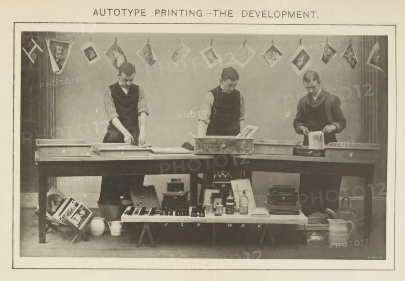 Process of developing what is believed to be a carbon print demonstrated by three men, Autotype Printing - The development, page, photomechanical print, inscription: 'I. Mounting (use of the Squeegee). 2. Developing Prints. 3. Fixing in Alum Solution. A. Cold Water Tray, No. IB Blotting Boards. C. Mounting Dish and Stool. D. Developing Tank. E. Cold Water Tray No. 2. F. Alum Bath G. Cold Water Tray No. 3. H. 'Hero' Stove. J. Johnson's Actinometers. R. Burton's Actinometers. S. Sawyer's Actinometers. T. Pressure Frames. P. Autotype Pressure Boxes. V. Packets of Cut Tissues. W. Waxing Compound. X. Collodion.', printmaker: Sawyer, printer (person): Autotype Company, The, c. 1885 - in or before 1895, Sawyer, Autotype Company, photographer, paper, collotype, photoprint, height 107 mm x width 175 mm, Print on a fold-out sheet opposite page 38., developing, film, A sepia photograph shows three men standing behind a long wooden table, each wearing dark aprons over shirts with rolled sleeves an...