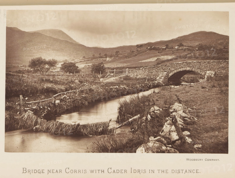 Bridge near Corris with Cader Idris in the distance, Bridge in a landscape near Corris, with a view of Cadair Idris in the distance, page, photomechanical print, Wales, printmaker: Woodbury Permanent Photographic Printing Company, c. 1881 - in or before 1891, photographer, paper, Woodburygravure, height 90 mm x width 141 mm, mountains, bridge, A sepia-toned landscape photograph showing a gently flowing stream bordered by grassy banks and scattered stones, a rustic wooden fence running alongside the water, a stone arch bridge crossing the stream, low rolling hills and terraced fields in the background, clusters of trees dotting the midground, and distant rounded mountains under a cloudy sky.