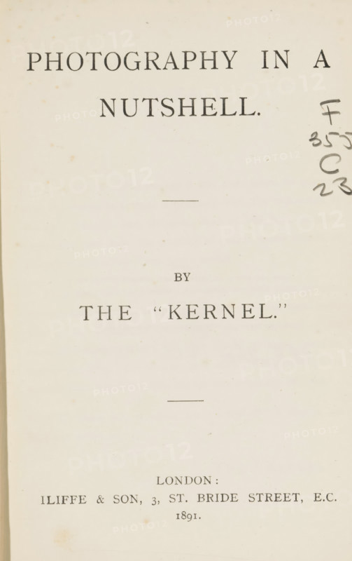 Photography in a nutshell, book, publisher: Iliffe & Son, London, 1891, textile materials, pasteboard, paper, linen (material), autotype, printing, Woodburygravure, kallitype, aututypie, Woodburygravure and callitype, height 189 mm x width 130 mm x thickness 20 mm, A pale, aged page with dark printed lines of large serif lettering across the top and center, a short horizontal rule near the upper middle and another near the lower middle, faint brown foxing spots scattered across the background, a column of dark handwritten ink marks along the right edge, and squared margins showing the inner binding edge on the left.