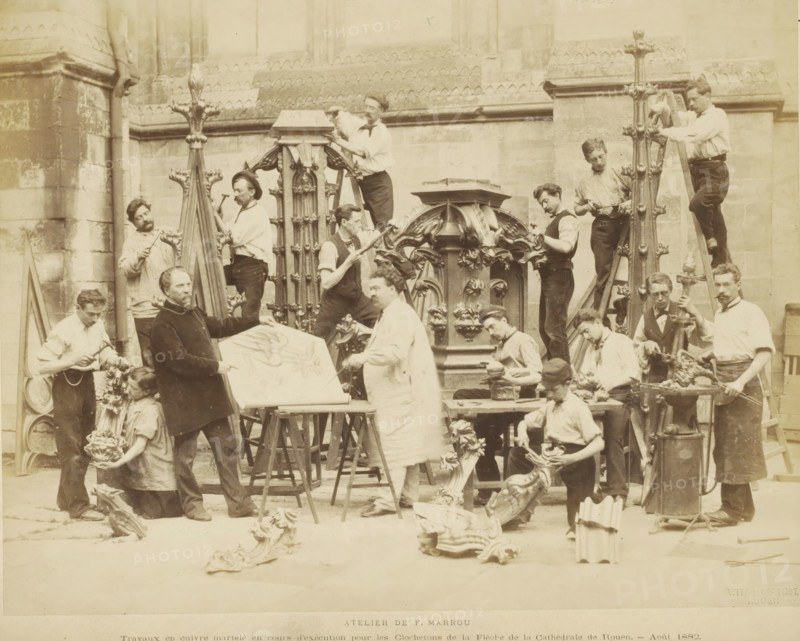 Atelier de F. Marrou Travaux en cuivre tortureé en cours d'exécution pur les Clochetons de la Flèche de la Cathédrale de Rouen., Sculptors at work for the studio of Ferdinand Marrou at Notre-Dame in Rouen, photograph, page, name: 'Witz & cie Phot. Rouen', photographer: Witz et Cie., Rouen, 1882-08, Witz et Cie., photographer, paper, albumen print, albumendruk, height 214 mm x width 273 mm, piece of sculpture, reproduction of a piece of sculpture, sculptor at work, A sepia-toned photograph shows a group of men and a woman working on ornate carved and cast decorative architectural pieces outside a stone building with buttresses and patterned masonry; several tall vertical carved finials and a central elaborate canopy-like structure are prominent, with men standing on ladders or holding tools while others shape or polish smaller decorative elements on tables and the ground; a few workers wear aprons or smocks and hold hammers, files, or clamps, while one man points at a large flat panel d...