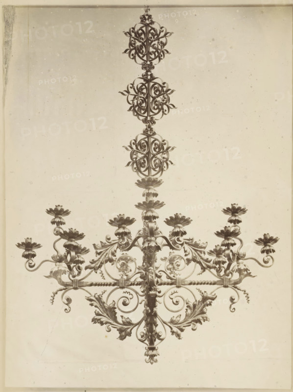 Chandelier decorated with leaf motifs, Luster en fer forgé et repoussé, photograph, page, name: 'Witz & cie Phot. Rouen', inscription: 'Composé et exécuté par F. Marrou, pour M. Depeaux à Rouen.', photographer: Witz et Cie., c. 1882 - in or before 1884, paper, albumen print, height 278 mm x width 215 mm, ornament derived from plant forms, chandelier, candelabrum, The image shows the upper portion of an ornate metal chandelier against a plain light background, featuring a central vertical rod with symmetrical scrollwork and decorative floral elements, multiple layered ornamental rosettes and leaf-like motifs, curled metal arms extending outward that support cup-shaped flower forms, and a repeating circular lattice of intricately twisted metalwork running horizontally across the piece.