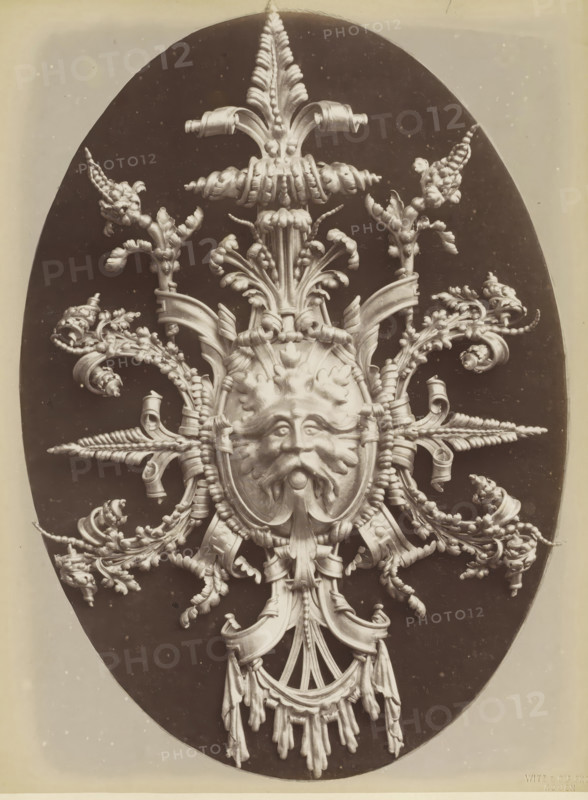 Decorative lead shield with a lion's head and leaf motifs, Cartonche en plomb repoussé an marteau, photograph, page, inscription: 'Composé et exécuté par F. Marrou.', name: 'Witz & cie Phot. Rouen', photographer: Witz et Cie., c. 1880 - in or before 1884, Witz et Cie., photographer, paper, albumen print, height 277 mm x width 208 mm, ornament derived from plant forms, lion's head, ornament, An ornate sculptural relief carved in a pale material is set against a dark oval background, featuring a central stylized human face with a mustache and flowing beard, surrounded by symmetrical arrangements of acanthus leaves, curling ribbons, braided elements, and botanical motifs that radiate outward. The composition includes scroll-like forms, twisted cords, and tassel-like hanging drapery beneath the face, with finely modeled textures and high-contrast lighting that emphasizes the three-dimensional details. The outer edges of the dark oval sit within a lighter rectangular border, and the overall...