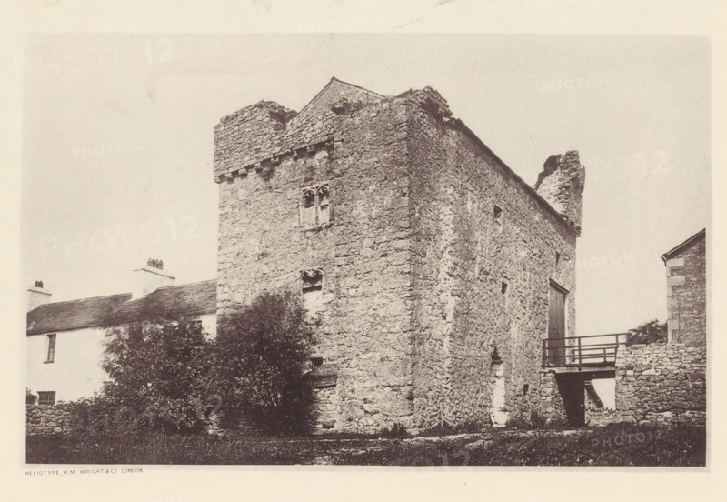 Wraysholme Tower, View of Wraysholme Tower, page, photomechanical print, photographer: Wright, HM & Co., Cumbria, London, c. 1870 - in or before 1880, paper, heliotype, height 111 mm x width 175 mm, tower, clock tower, monumental adornment, A sepia-toned photograph of a stone building with rough textured walls and uneven masonry, showing a tall rectangular tower-like section with small arched window openings and a crenellated top; a pitched roof and a ruined chimney or jagged stone projection rise behind it, while a wooden balcony or walkway with railings connects to the side where a doorway is visible; in front of and beside the building there are bushes and a low stone wall, and adjacent to the main structure a smaller whitewashed house with a sloped roof and chimneys is partially visible.