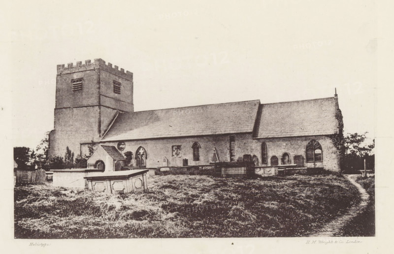 Urswick church, View of Saint Mary and Saint Michael's Church in Urswick, page, photomechanical print, photographer: Wright, HM & Co., Urswick, cliché maker: Wright, London, c. 1870 - in or before 1880, Wright, paper, heliotype, height 134 mm x width 187 mm, church (exterior), Cumbria, Saint Mary and Saint Michael's Church, A sepia-toned photograph of a stone church with a square bell tower topped by battlements on the left and a long nave with a pitched roof extending to the right, showing multiple arched windows along the side and a small entrance structure with a gabled roof in front of the tower; a grassy churchyard with scattered gravestones, a raised stone tomb in the foreground, and a low fence or enclosure near the building; trees and shrubbery appear behind and beside the church, and the sky above is light and empty.