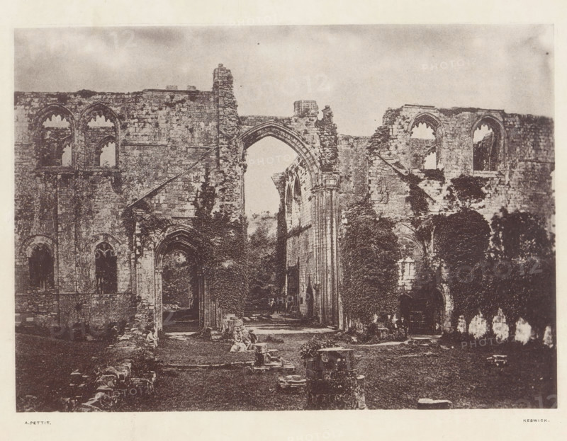 Furness Abbey_the church, View of the ruins of Furness Abbey, page, photomechanical print, photographer: Alfred Pettitt, Furness Abbey, Keswick, c. 1870 - in or before 1880, paper, heliotype, height 118 mm x width 166 mm, abbey, monastery, convent, Roman Catholic church, Cumbria, A wide view of a stone ruin with tall arched openings and fragmented walls, showing gothic-style window arches and doorways, crumbling masonry with exposed blocks and weathered surfaces, a large central archway leading into an interior space framed by tall columns and remnants of vaulted structures, areas of vegetation and ivy growing on and around the stonework, scattered fallen stones and rubble in the foreground, and a pale, cloudy sky visible above the ruined walls.