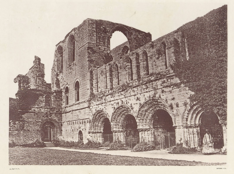 View of the courtyard of the ruins of Furness Abbey, Furness Abbey_the cloisters court, page, photomechanical print, photographer: Alfred Pettitt, Furness Abbey, Keswick, c. 1870 - in or before 1880, paper, heliotype, height 127 mm x width 172 mm, courtyard, abbey, monastery, convent, Roman Catholic church, Cumbria, A sepia-toned photograph shows a large stone building with a series of arches and tall narrow windows, some arches forming doorways and others set higher in the wall; the masonry appears weathered and textured, with sections of ruined upper walls and an opening where a large arch is partially intact; vegetation climbs part of the facade and low shrubs grow at the base, while a paved path runs along the front and an expanse of grass is visible in the foreground; a fragment of a statue or carved figure sits near one corner of the building.