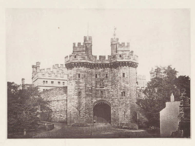 Lancaster Castle. 1876, View of Lancaster Castle, page, photomechanical print, photographer: Wright, HM & Co., Lancaster (Lancashire), London, 1876, paper, photoaquatint, height 127 mm x width 180 mm, castle, gate, entrance, Lancashire, Lancaster Castle, A stone gatehouse with battlements and two projecting round towers frames a large arched entrance, with masonry showing visible blocks and small slit windows; decorative corbels run beneath the parapet and a carved emblem is set above the arch; surrounding buildings with crenellated walls and rectangular windows rise behind the gatehouse; mature trees flank the structure and a curved stone wall and railing lead toward the entrance; a cobbled forecourt and a solitary lamp post stand before the arch, and a partial ruined wall appears at the right edge.