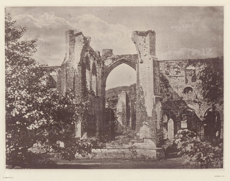 Furness Abbey_the east window, View of the east window of the ruins of Furness Abbey, page, photomechanical print, photographer: Alfred Pettitt, Furness Abbey, Keswick, c. 1870 - in or before 1880, paper, heliotype, height 125 mm x width 170 mm, abbey, monastery, convent, Roman Catholic church, church-window, eg rose-window, Cumbria, A sepia-toned photograph showing the stone remains of a large ruined building with tall arched openings and broken columns, parts of the walls covered in climbing vegetation, leafy trees and bushes framing the left and right sides, a cloudy sky above, and a low stone platform in the foreground.