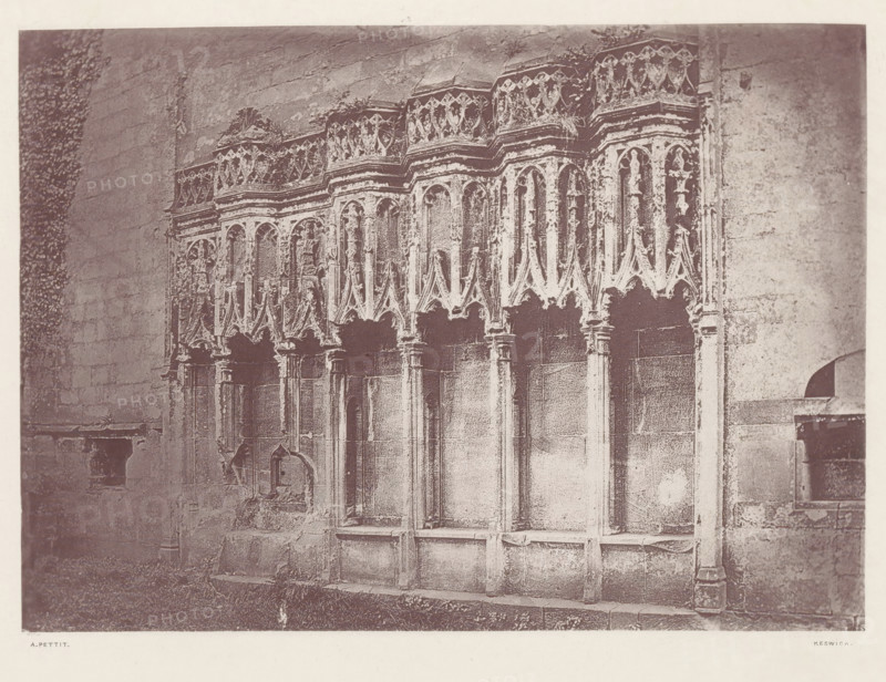 Furness Abbey_the sedilia, Sedilia in the ruins of Furness Abbey, page, photomechanical print, photographer: Alfred Pettitt, Furness Abbey, Keswick, c. 1870 - in or before 1880, paper, heliotype, height 125 mm x width 175 mm, abbey, monastery, convent, Roman Catholic church, parts of church interior, Cumbria, A section of an ornate stone wall with a series of tall, narrow arched niches topped by decorative carved tracery and pinnacles, supported by slender columns and detailed capitals, with weathered surfaces and patches of discoloration; a rough stone floor and a strip of grass run along the base; portions of adjacent plain stone walls frame the carved section on both sides.