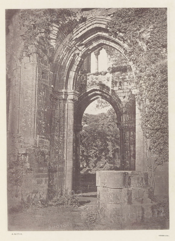 Arch at Furness Abbey, Arch of the ruins of Furness Abbey, page, photomechanical print, photographer: Alfred Pettitt, Furness Abbey, Keswick, c. 1870 - in or before 1880, paper, heliotype, height 169 mm x width 123 mm, arch, archivolt, architecture, ruin of church, monastery, Cumbria, A tall stone archway with layered carved voussoirs frames an opening to a leafy scene beyond, flanked by massive square columns and textured masonry walls showing weathering and patches of vegetation; remnants of another arch and column are visible in front, and climbing plants cover parts of the right wall while rough stone blocks and low grassy ground occupy the foreground.