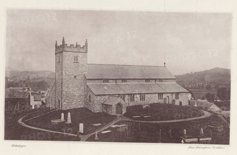 View of Saint Michael and All Angels Church in Hawkshead, Hawkshead church, page, photomechanical print, photographer: Alex Rivington, Hawkshead, London, c. 1870 - in or before 1880, paper, heliotype, height 107 mm x width 181 mm, church (exterior), Cumbria, Saint Michael and All Angels Church, A stone church with a square tower and crenellated top stands on a grassy hill, its long pitched roof and stone walls visible along with a small arched porch entrance and several narrow windows. A curving path winds through a churchyard containing various headstones and tombs of different shapes and sizes, some enclosed by low railings. In the background there are rolling hills and scattered trees, and adjacent to the church a cluster of small buildings and rooftops can be seen. The image has a sepia tone and an overall aged photographic appearance.